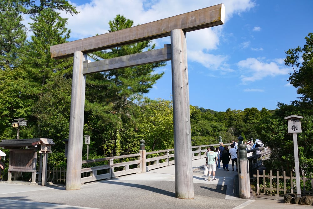 A large wooden torii gate stands at the entrance to a bridge leading to a forested area, with several people walking across.