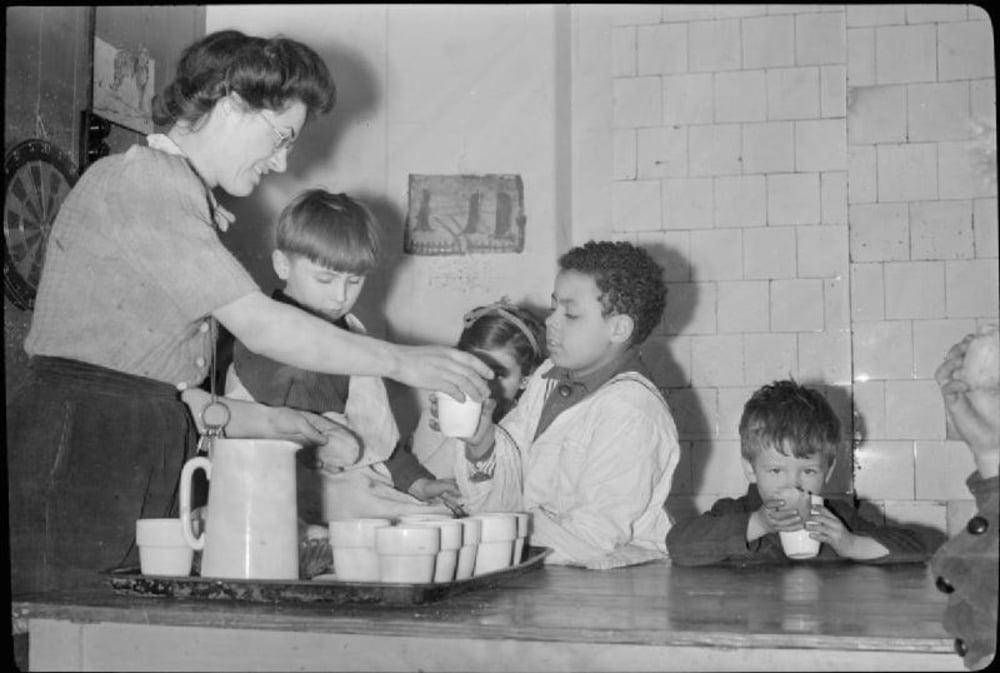 A woman serves cups of milk to a group of young children sitting at a table.