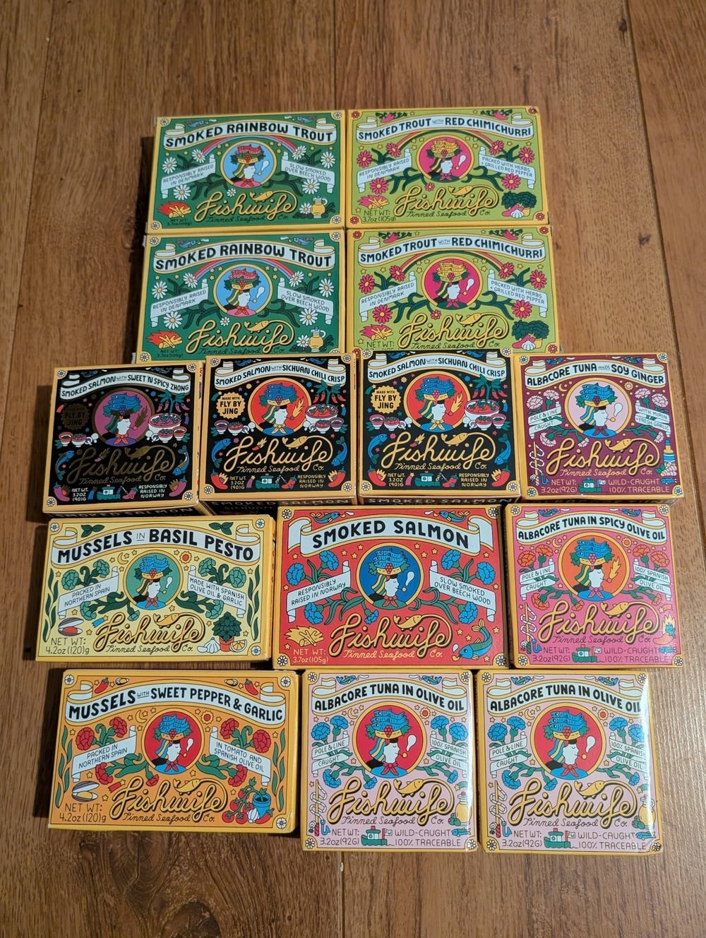 Fourteen colorful tins of Fishwife tinned seafood arranged in a pyramid on a wooden surface, including varieties like Smoked Rainbow Trout, Smoked Salmon, Mussels, and Albacore Tuna.