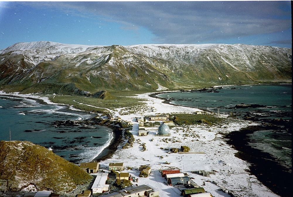 An aerial view of a research station with small buildings and a geodesic dome located on a snow-dusted isthmus between two bodies of water, backed by a large, snow-capped mountain range.