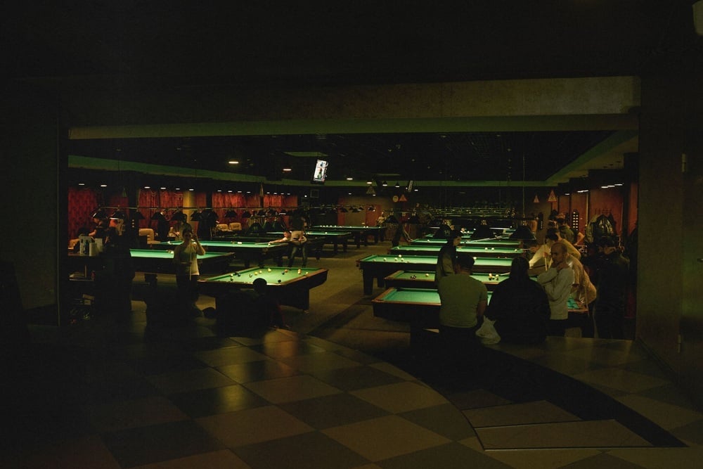 People playing billiards in a dimly lit pool hall filled with multiple tables.
