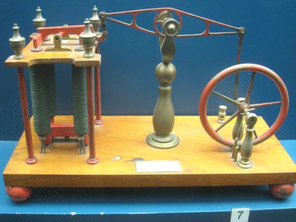 A historical scientific instrument featuring two large electromagnetic coils connected by a metal arm to a manual flywheel on a wooden base.
