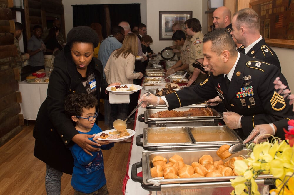 A soldier in uniform serves gravy onto a plate held by a young boy, as a woman guides his hand at a buffet line.