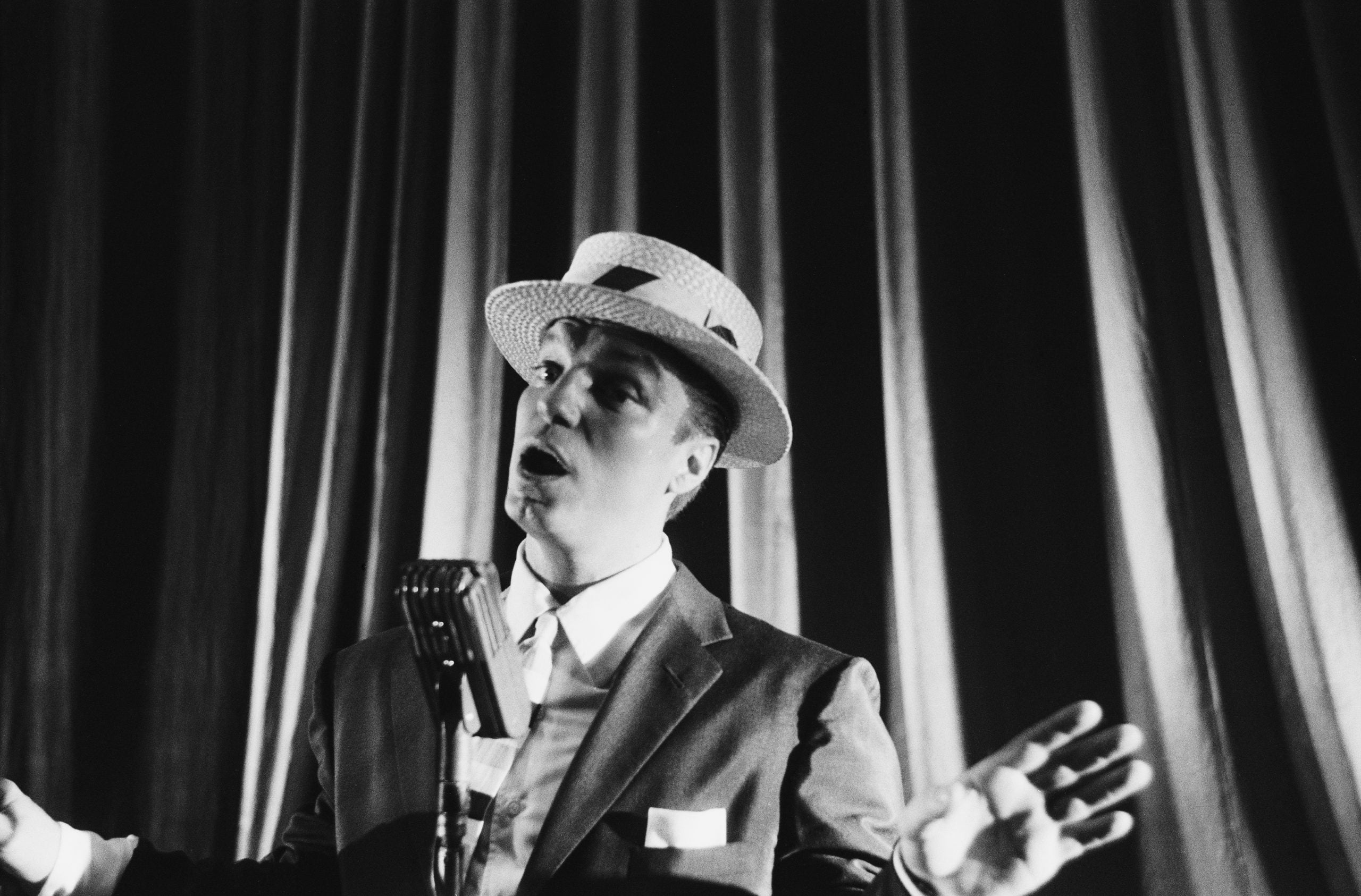 A man in a straw hat and suit jacket singing into a vintage microphone in front of curtains.