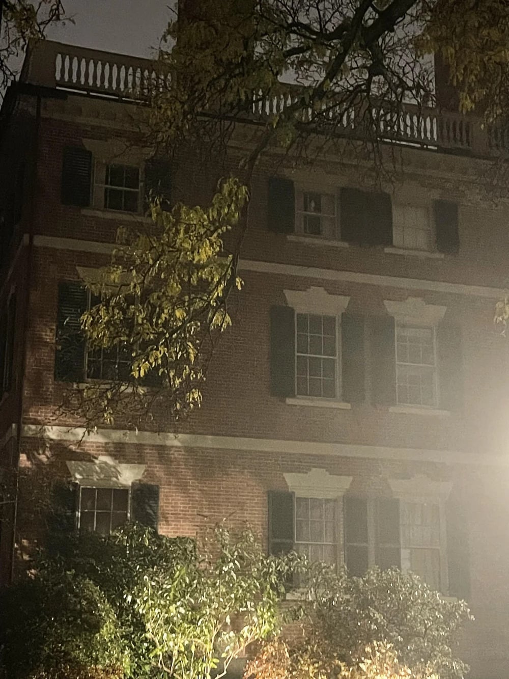 A three-story brick building with a balustrade roof at night, illuminated by soft light through surrounding trees.