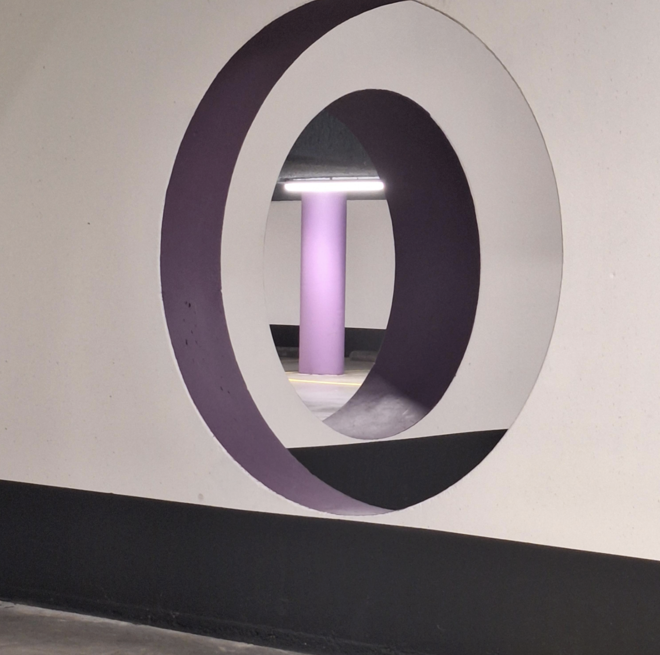 A purple-rimmed circular opening in a white wall looking through to a parking garage with a purple support pillar.