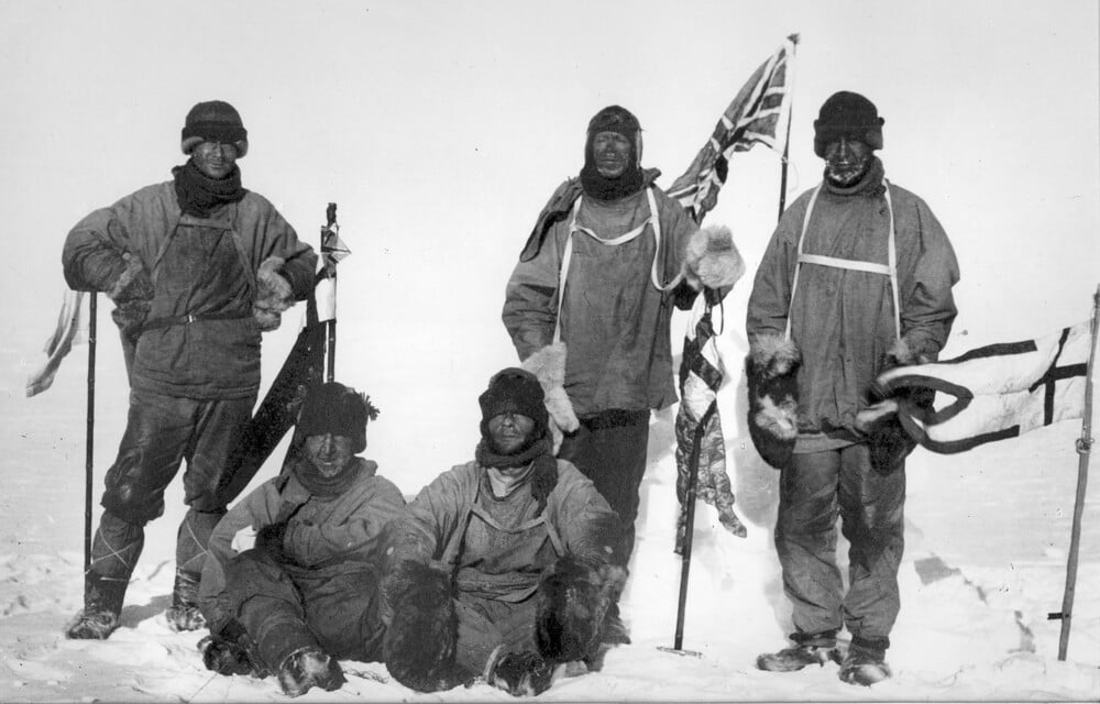 Five explorers in winter gear pose in the snow with a British flag.