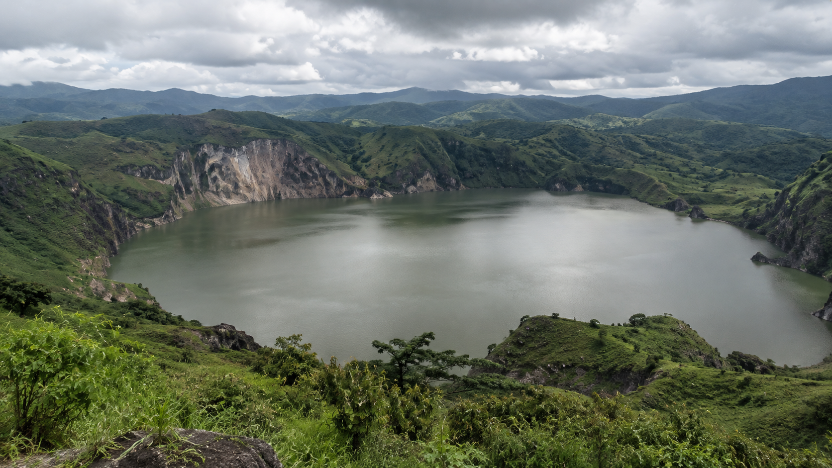 A large, circular crater lake surrounded by lush green hills under a cloudy sky.