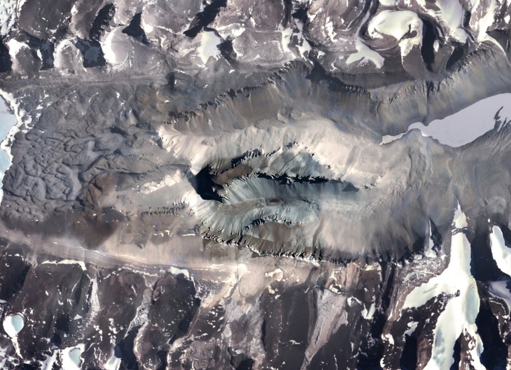 Satellite view of a dark, crater-like mountain formation surrounded by snow and glacial ice.