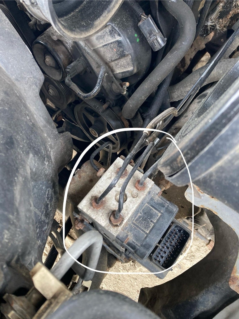 Close-up of a vehicle’s engine bay, with a white circle highlighting the ABS hydraulic control unit and its connected wiring harness.