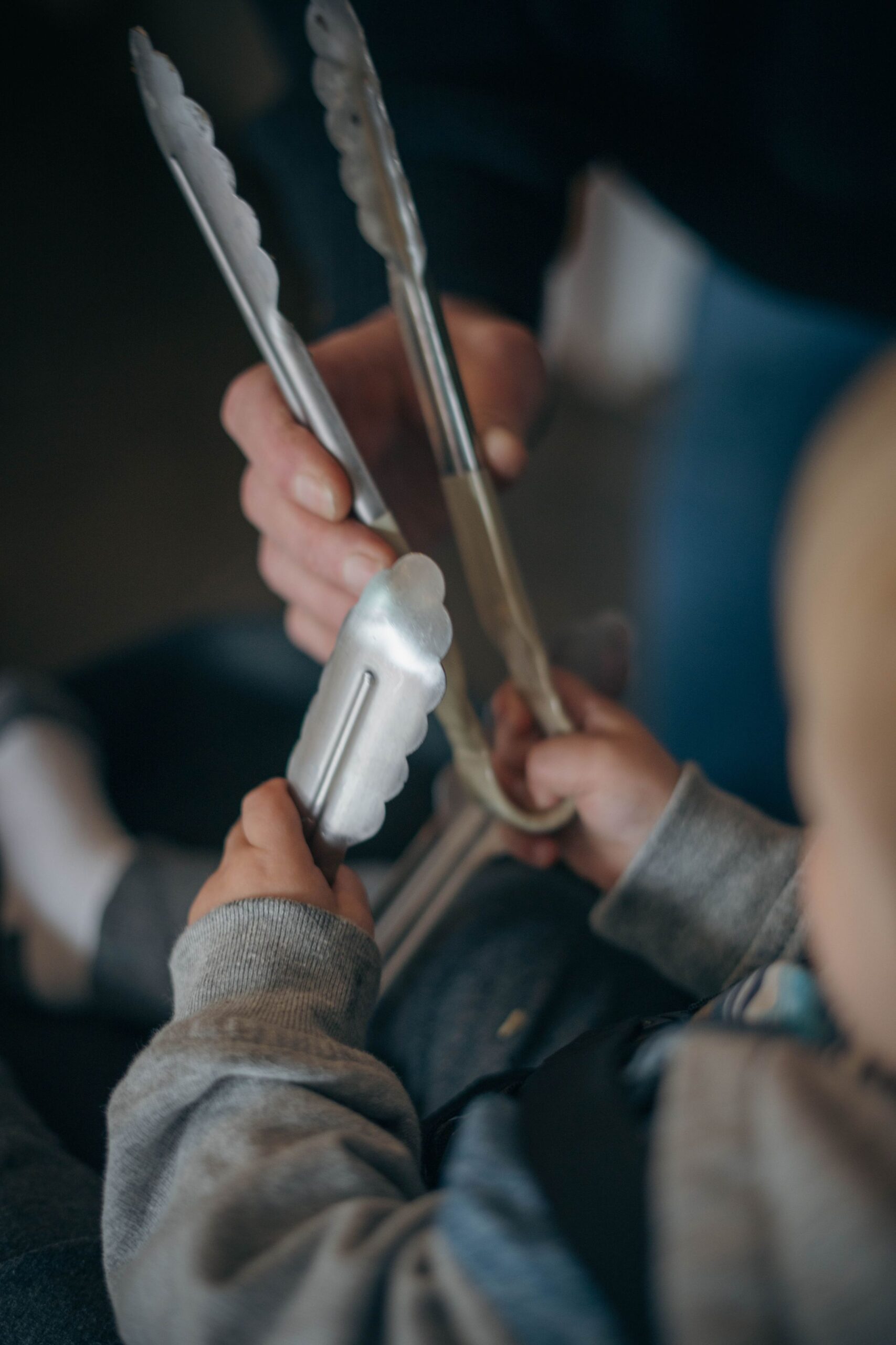 A child and an adult each hold metal kitchen tongs, facing each other. The focus is on their hands, with the background softly blurred.