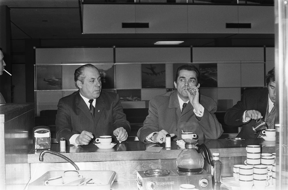 Three men in suits sit at a counter drinking coffee; two are smoking cigarettes, and one is pouring milk. The scene appears to be in an airport cafe, with airplane images visible on panels behind them.