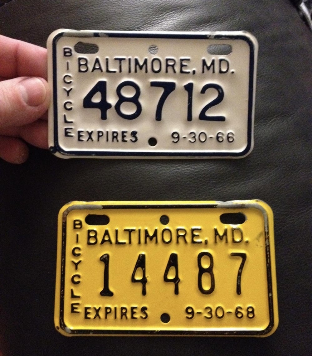Two vintage Baltimore, Maryland bicycle license plates: a white plate numbered 48712 expiring 9-30-66, and a yellow plate numbered 14487 expiring 9-30-68.