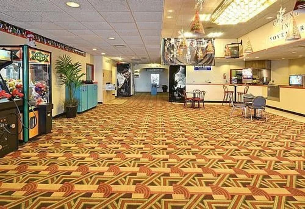 A brightly lit, wide indoor hallway with patterned carpet, featuring a claw machine, a dining area with tables and chairs, and decorative posters on the walls.