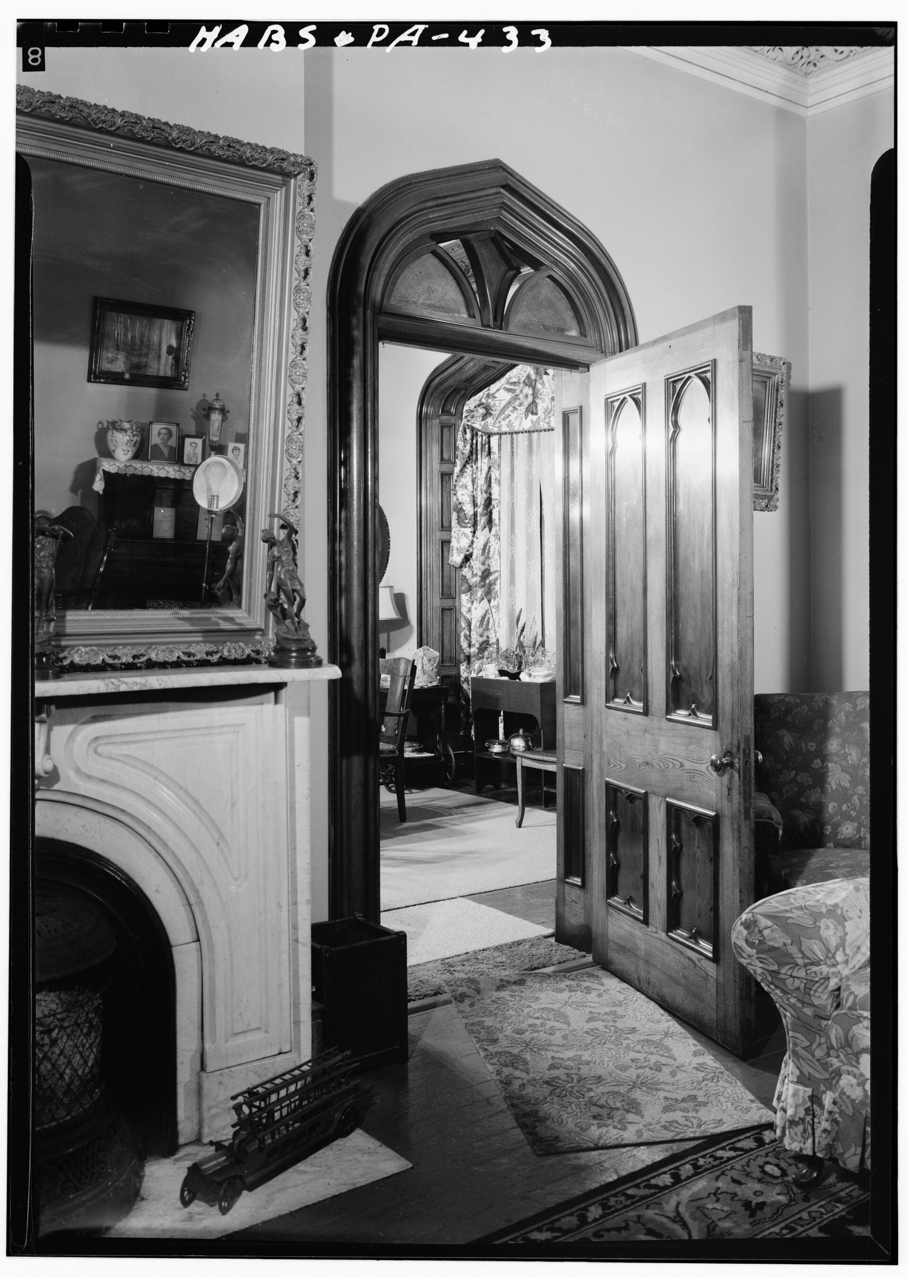 Black and white photo of a room featuring a marble fireplace with a large mirror, a gothic-arched doorway opening to an adjacent room, and patterned upholstered furniture.