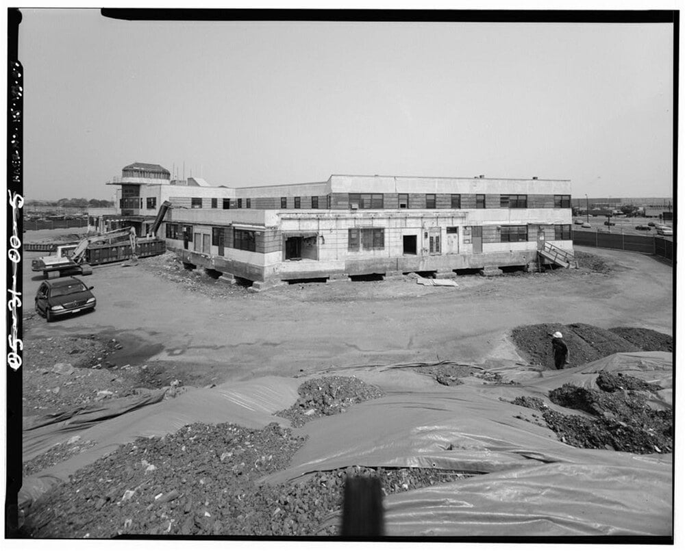 A low-angle, black-and-white view of a multi-story building under renovation, with construction machinery nearby and a person in a hard hat standing in a dirt-covered foreground.