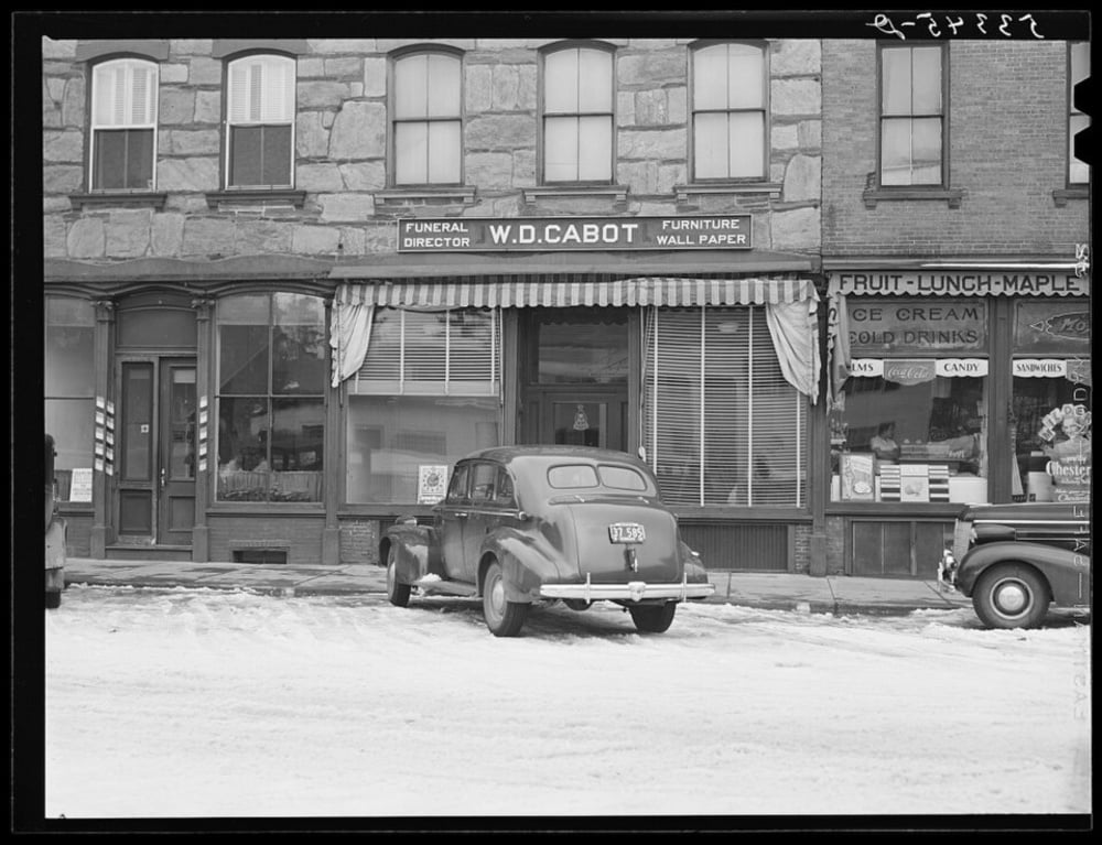 A vintage car parked on a snowy street in front of W.D. Cabot funeral home and a neighboring shop advertising fruit, lunch, and ice cream.