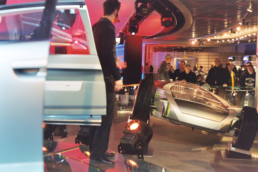 A presenter stands next to a car chassis on display at an indoor exhibition while a small group of people watches.