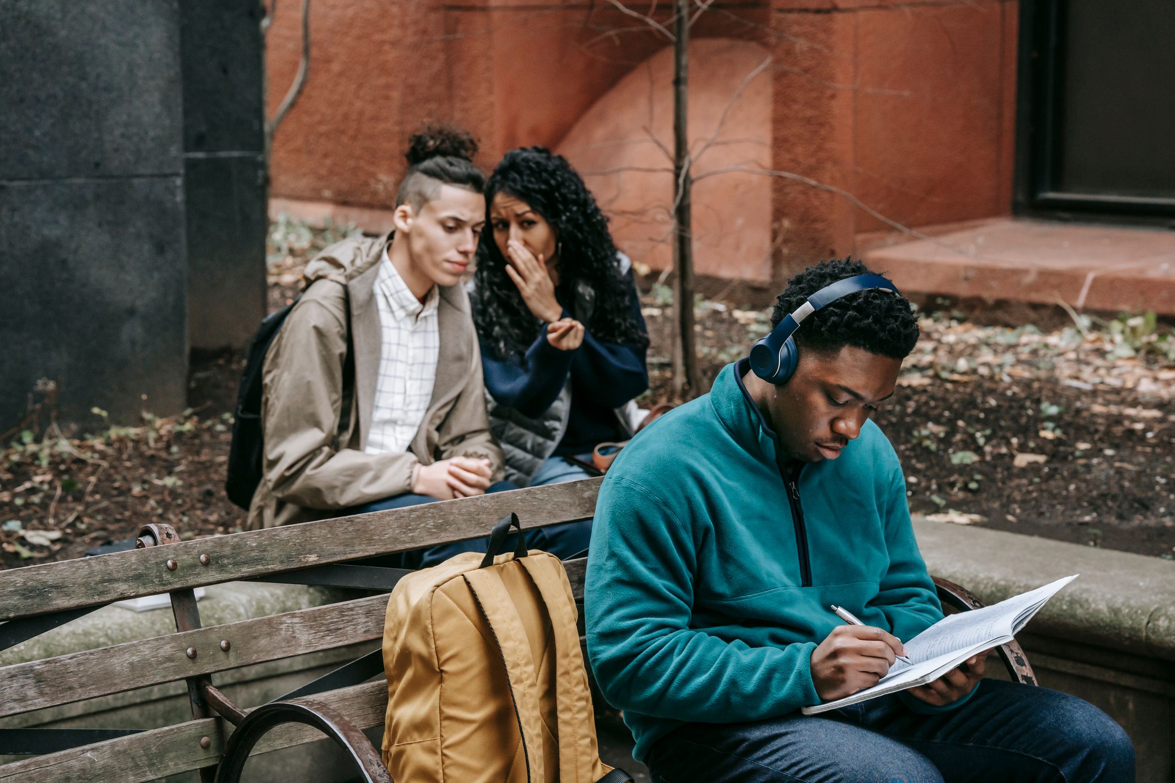 A man wearing headphones writes in a notebook on a bench, while two people behind him whisper to each other, one cupping their hand over their mouth. A tan backpack sits on the bench.