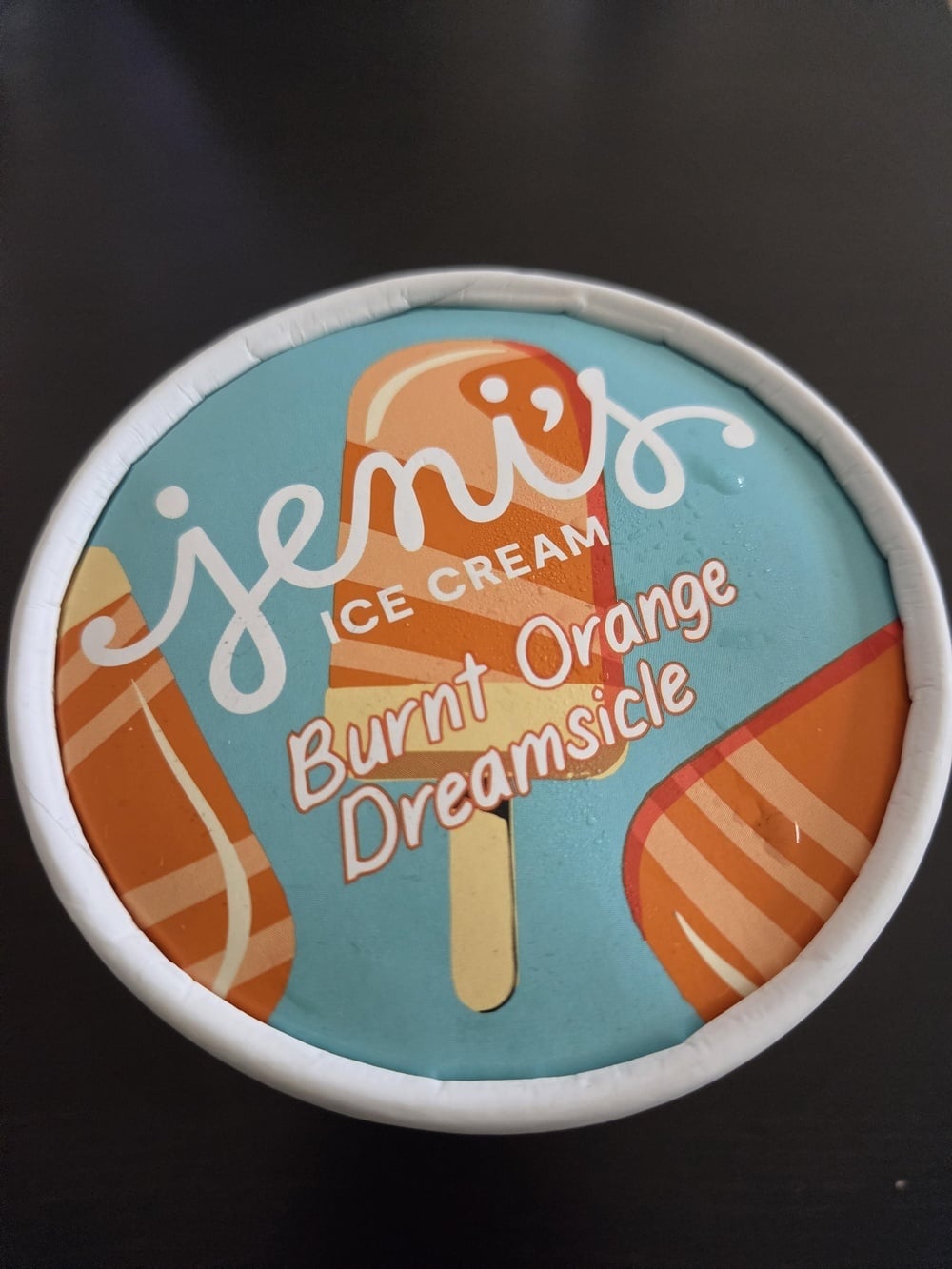 A round pint lid of Jeni’s Burnt Orange Dreamsicle ice cream featuring an illustration of an orange popsicle.