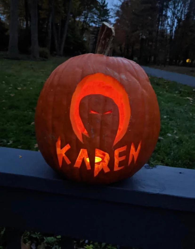 A carved jack-o’-lantern sitting on a railing, featuring a silhouette of a person in a hood above the word “KAREN” cut into the pumpkin’s face.