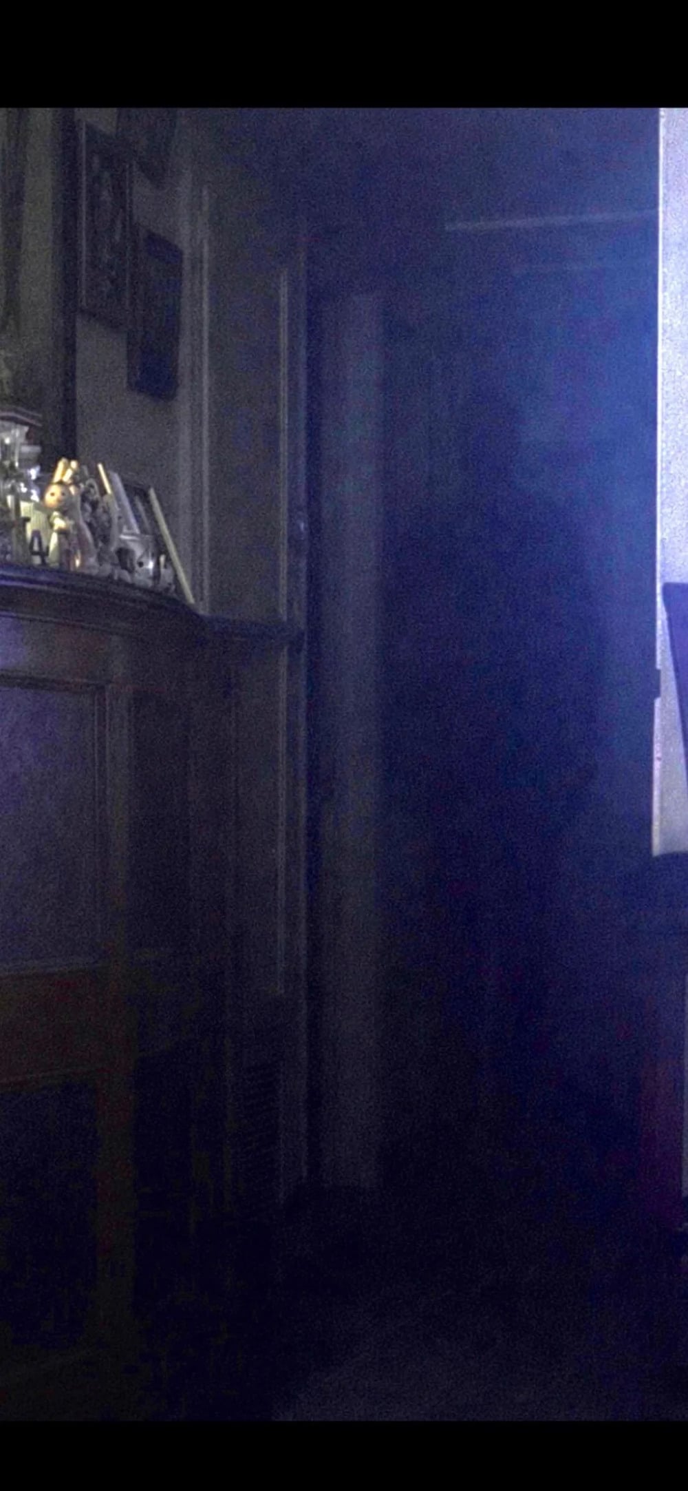 A dark, shadowy human-like silhouette stands in a dimly lit doorway next to a wooden cabinet.