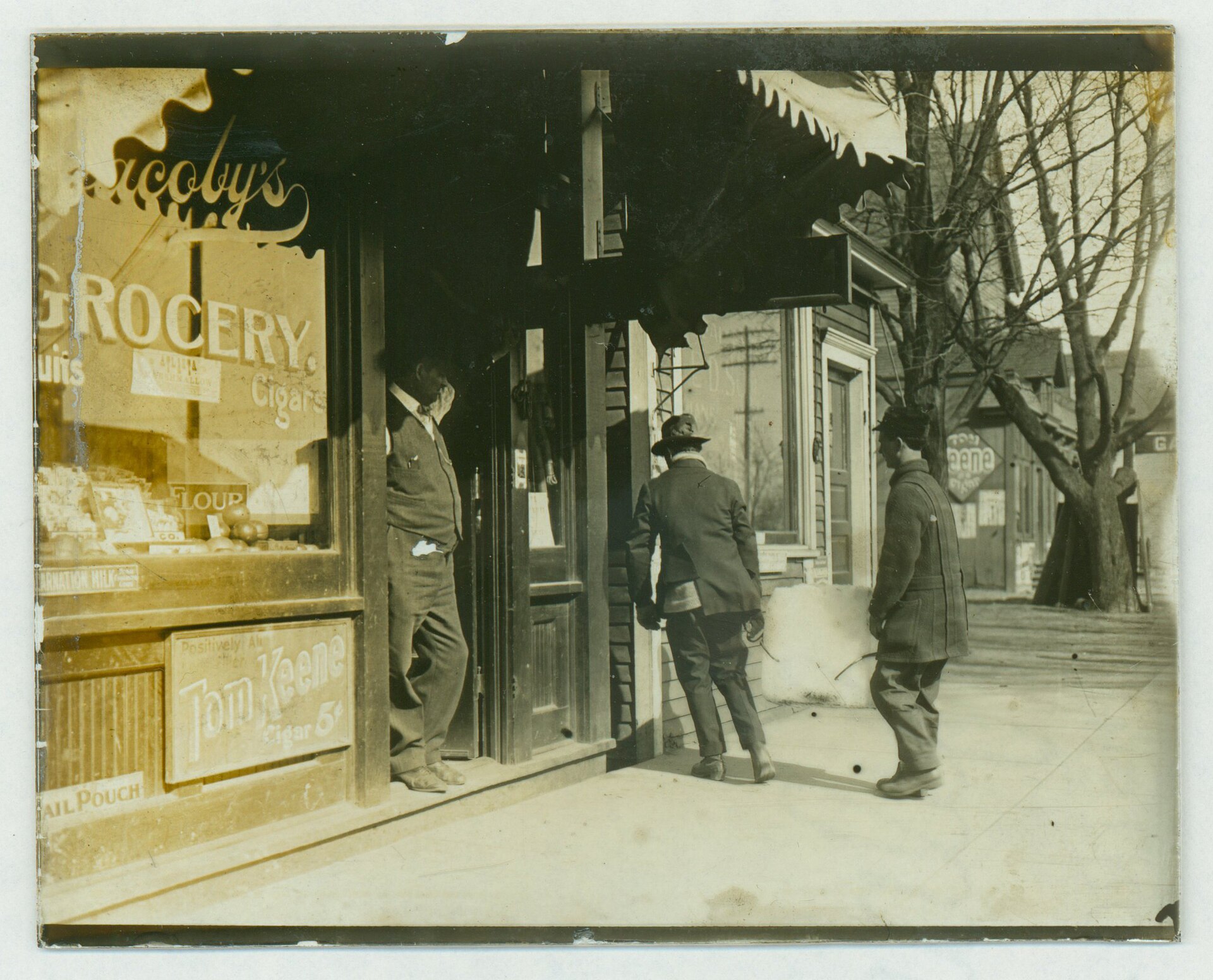 A vintage street scene showing a man standing in the doorway of a grocery store while two other men walk by on the sidewalk.