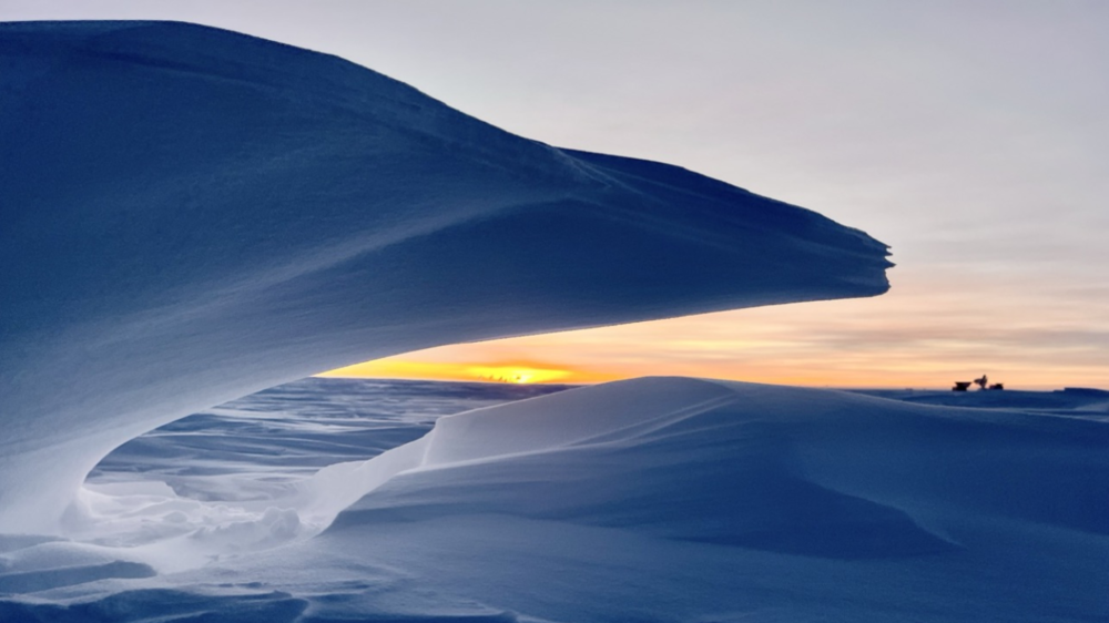A large, overhanging snowdrift in the foreground with a sunset visible on the horizon.