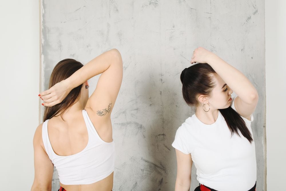 Two women in white tops stand in front of a gray wall, each with one arm raised and the other hand holding their head, appearing to stretch their necks and shoulders.