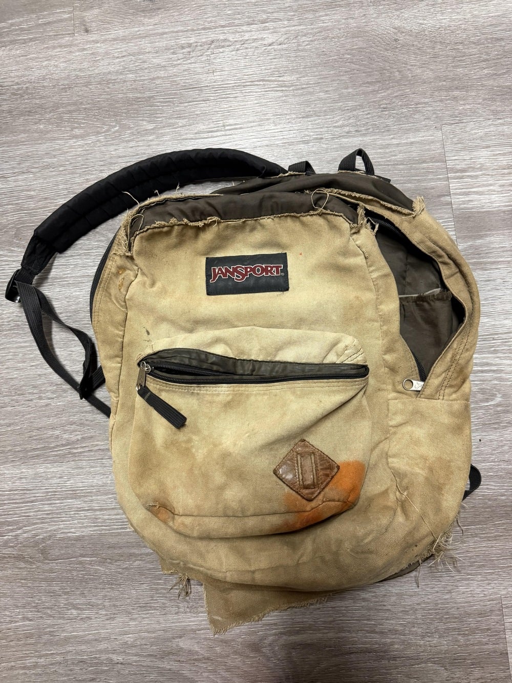 A worn and dirty tan Jansport backpack with frayed edges and a large orange stain on the front pocket sits on a wooden floor.