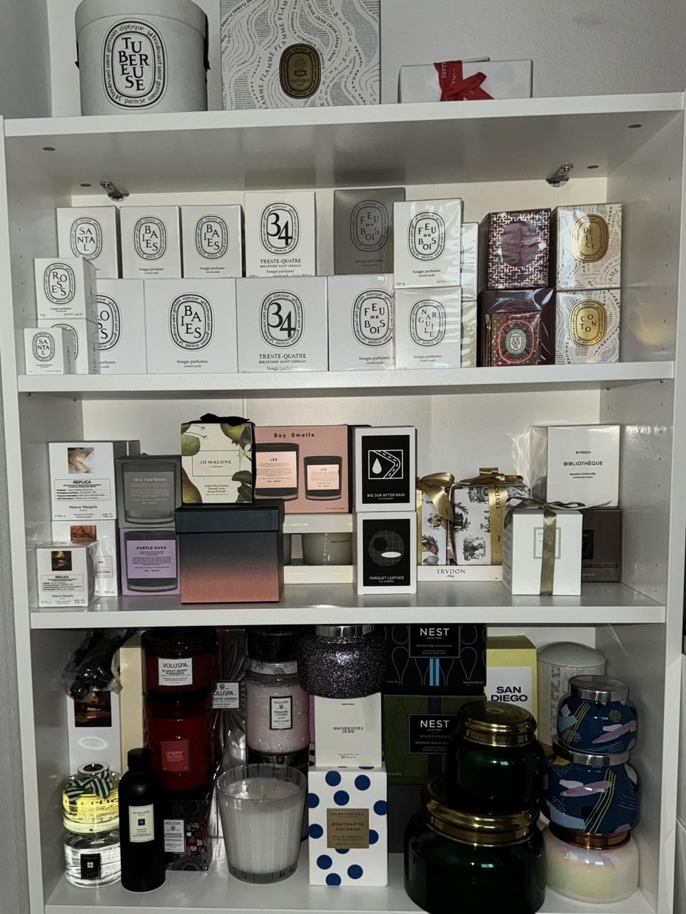 A white shelving unit filled with various luxury scented candles and fragrance boxes from brands including Diptyque, Jo Malone, Boy Smells, Trudon, and Nest.
