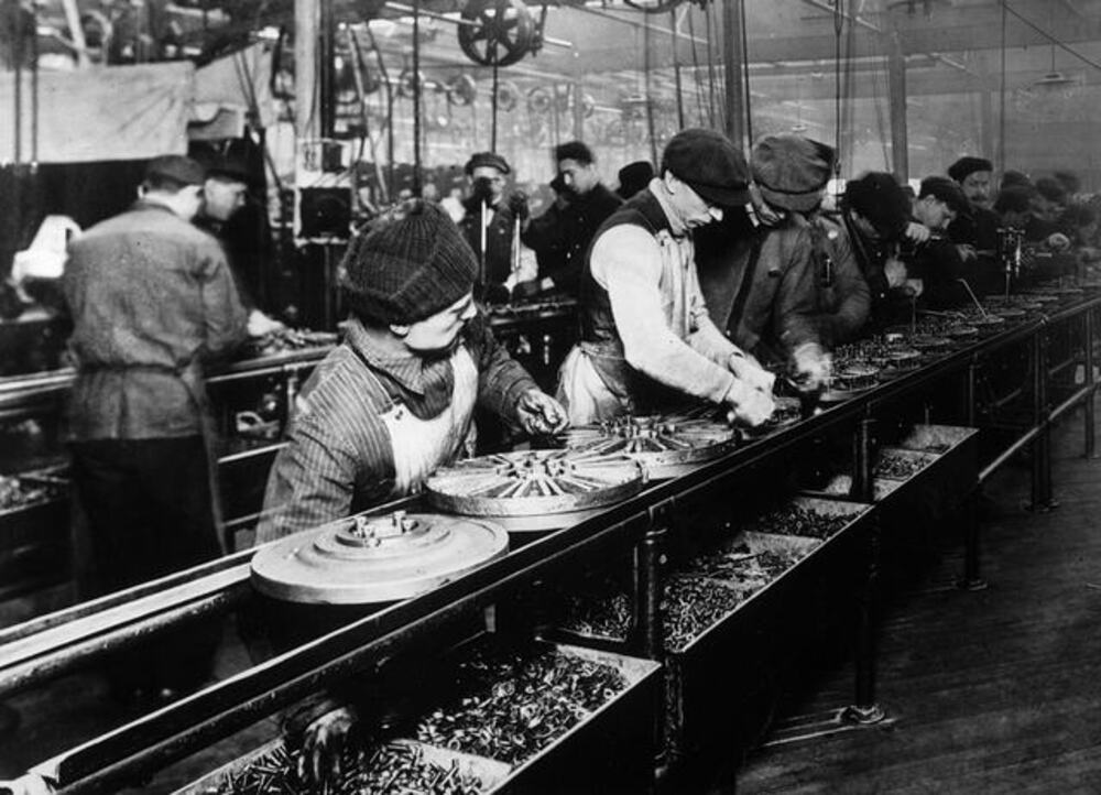 A line of workers in caps and aprons assemble metal parts on a factory conveyor belt.