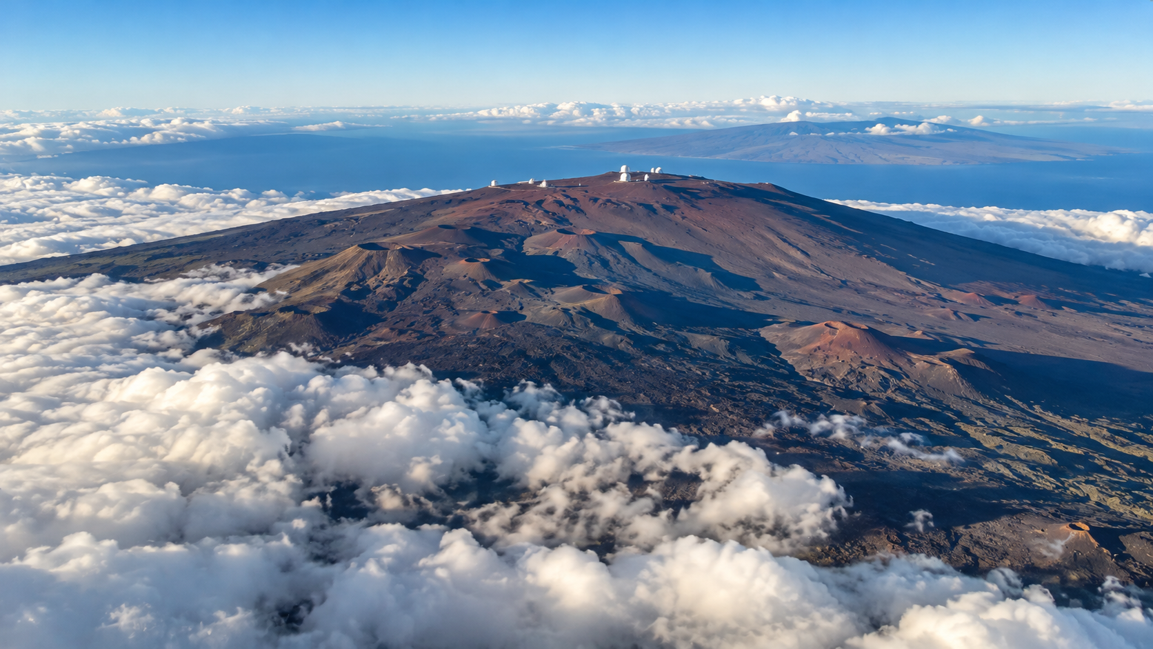 Aerial view of the Mauna Kea summit with its observatories rising above a layer of clouds, with another island visible in the distance.
