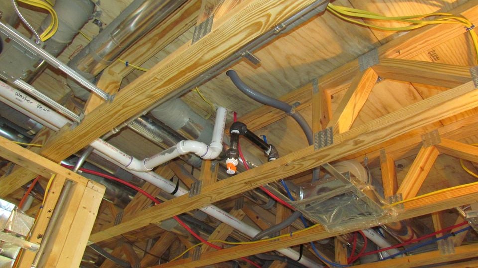 Exposed ceiling joists in unfinished construction showing plumbing pipes, electrical wiring, and HVAC ducting.