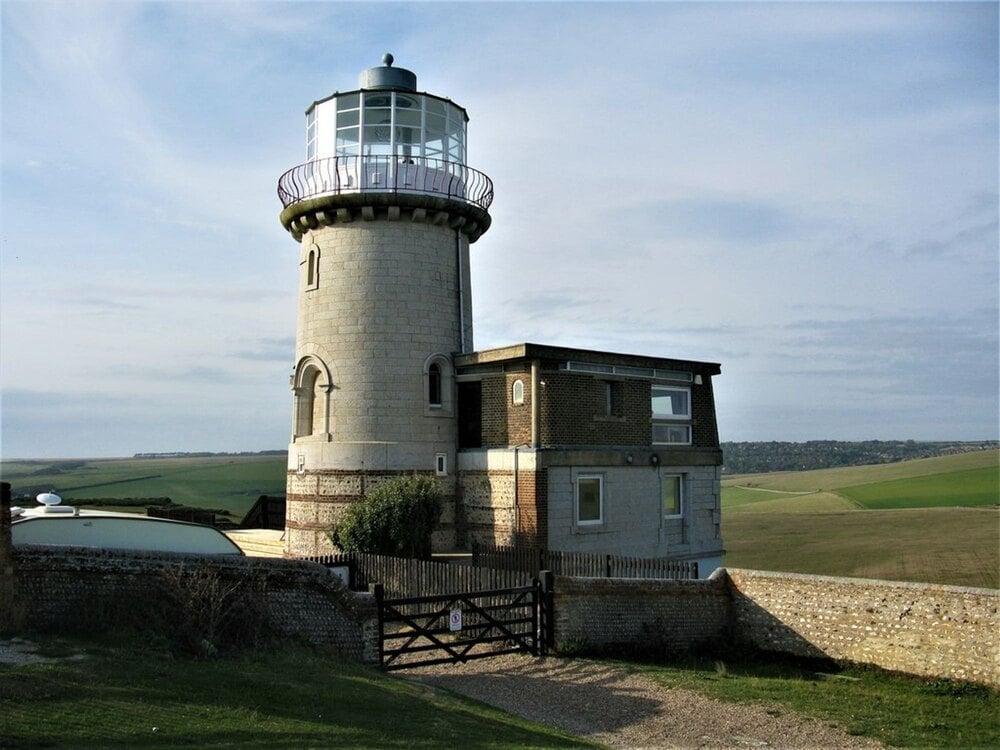 A lighthouse attached to a brick residential building, surrounded by a low stone wall and a grassy landscape.