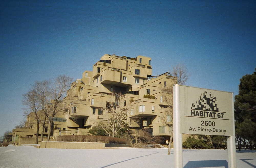 The Habitat 67 housing complex, a modular structure of stacked concrete cubes, seen behind a sign identifying the site at 2600 Av. Pierre-Dupuy in a snow-covered landscape.