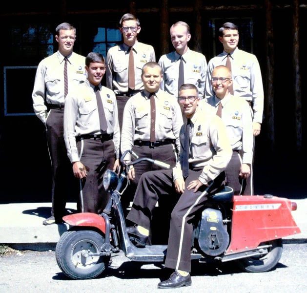Seven men in uniforms posing with a red Cushman motor scooter in front of a building.