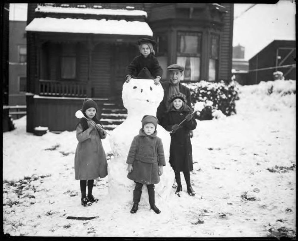 A man and four children pose with a snowman in a snowy yard in front of a house.