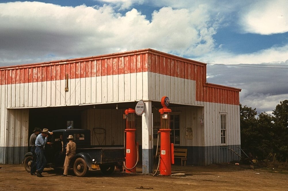 Three men stand beside a vintage pickup truck at a roadside gas station with red, white, and blue siding.