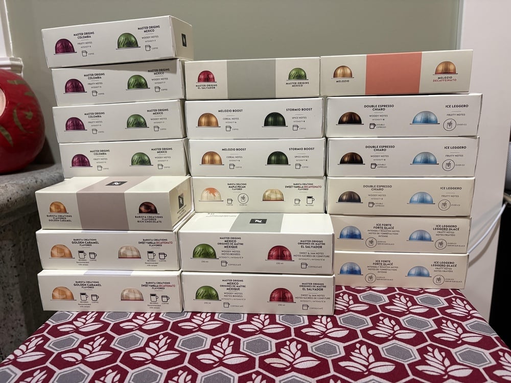 A stack of twenty-five Nespresso coffee capsule boxes in various colors, flavors, and intensity levels arranged on a patterned tablecloth.