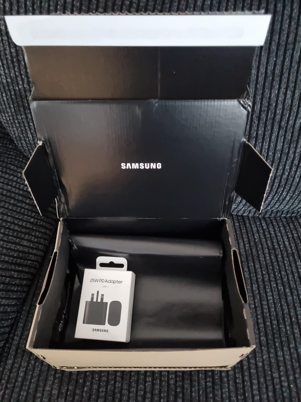 An open Samsung box sits on a striped fabric surface. Inside the box is a white package labeled “25W PD Adapter” with an image of a charging adapter and cable. The box interior is black with “SAMSUNG” printed inside the lid.