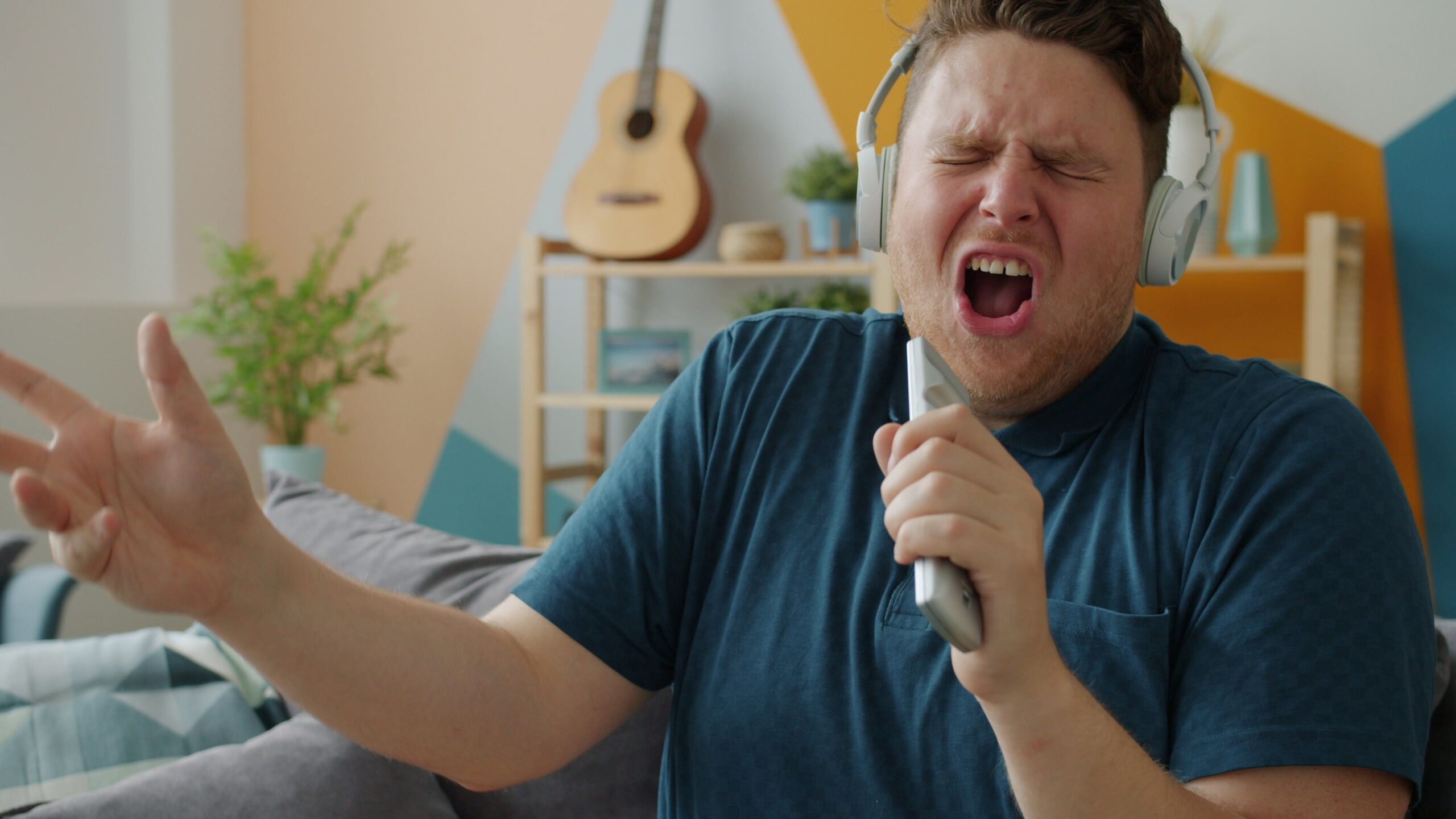 A man wearing headphones passionately sings into a TV remote in a living room. A guitar hangs on the wall behind him, and a plant sits on a shelf.