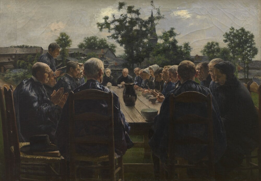 A group of people seated around a long outdoor table, their hands clasped in prayer before a meal.