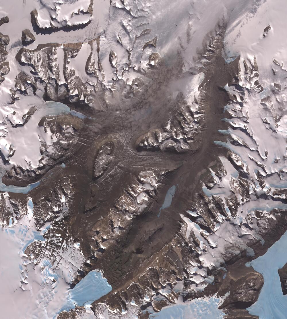 Satellite view of a mountainous landscape with snow-covered peaks, dark rocky ridges, and glaciers.