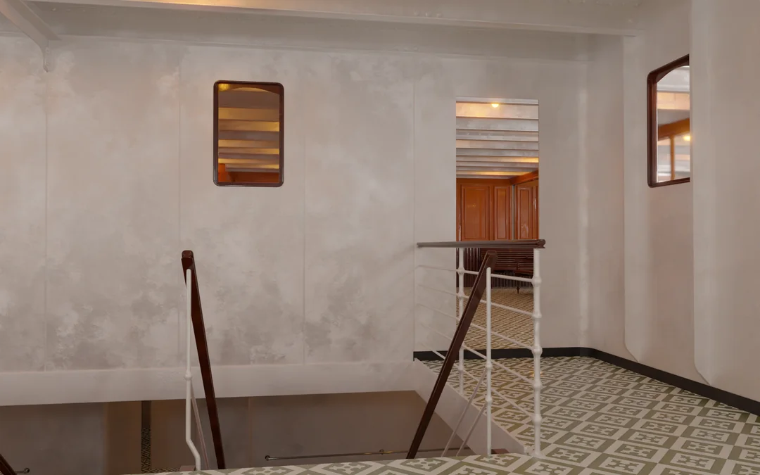 A staircase with white metal railings and dark wooden handrails leads to a lower deck, viewed from a hallway with a patterned tile floor and two framed mirrors on the wall.