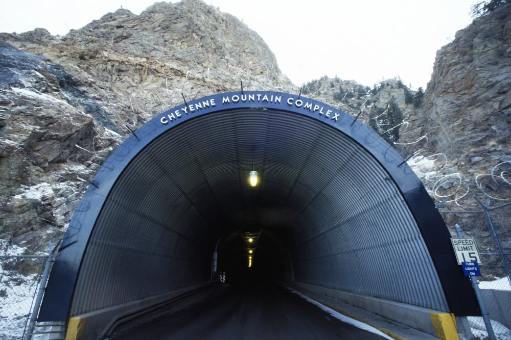 The arched metal entrance to the Cheyenne Mountain Complex tunnel, built into a rocky mountainside with a speed limit sign nearby.