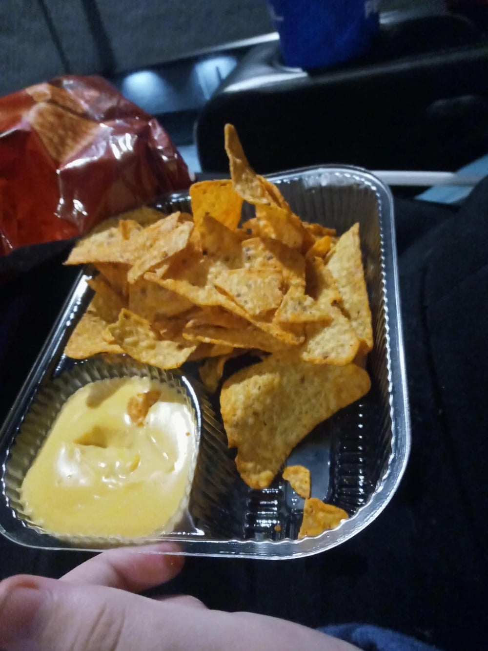 A plastic container of tortilla chips and a side of cheese sauce, held by a person.
