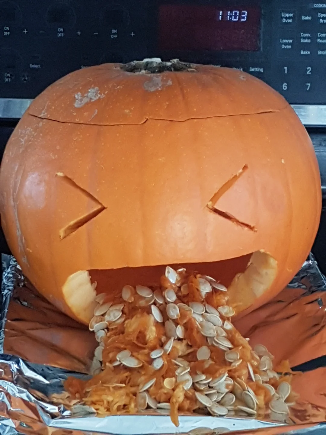 A carved pumpkin with a wide-open mouth, filled with scooped-out pumpkin pulp and seeds spilling onto foil.