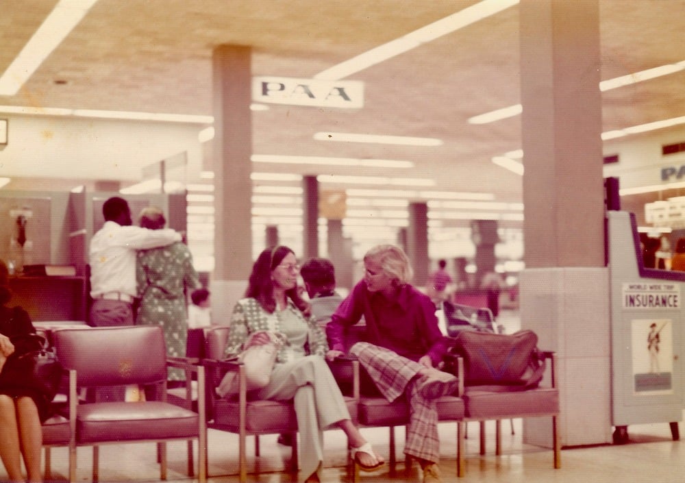 Two people sit and talk in an airport waiting area with bags at their feet. Other travelers stand and walk nearby. A sign reads “PAA,” and another sign advertises insurance. The scene appears to be from the 1970s.
