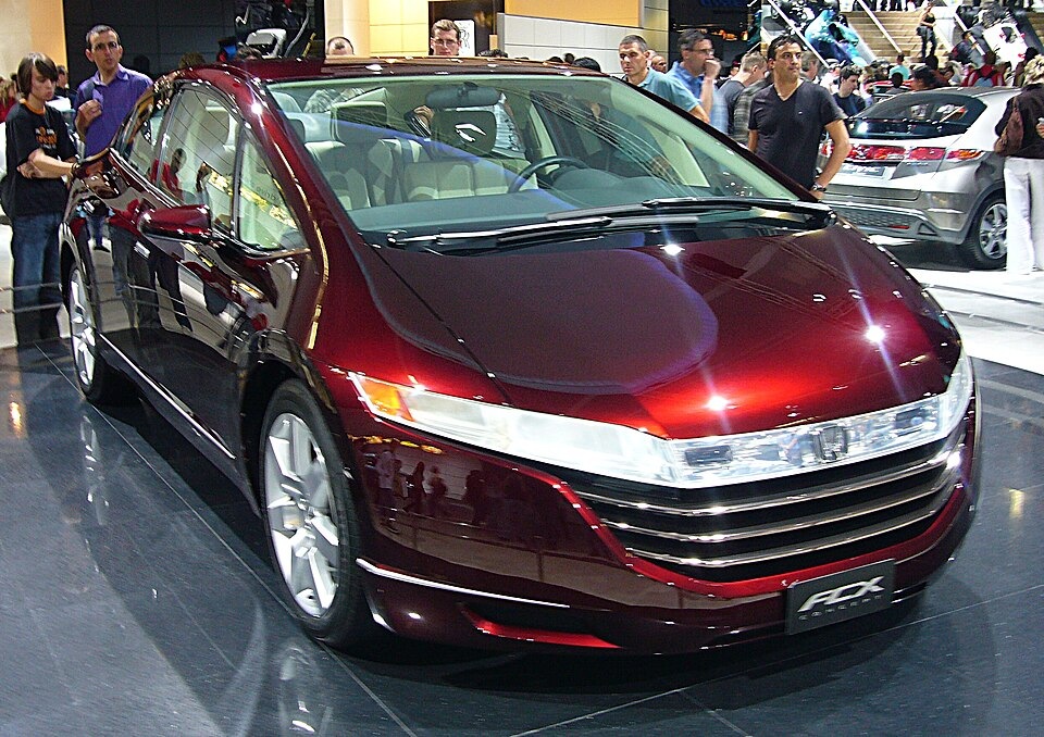 A dark red Honda FCX Concept car displayed on a showroom floor surrounded by people.